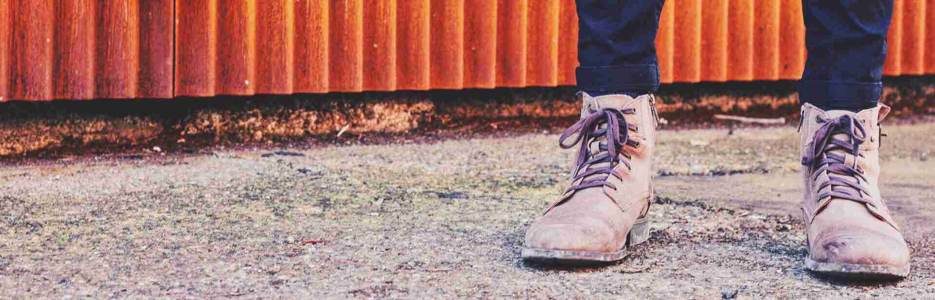 2 legs in jeans with dusty working boots on a gravel trail in front of an orange rusty wavey wall. The picture is cut vertically before the knees.